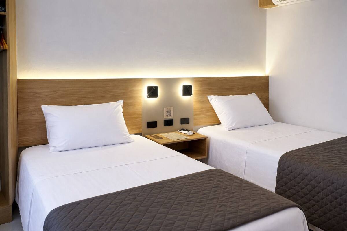 A hotel room with two single beds, a wooden headboard, bedside tables with lamps, and a small nightstand.