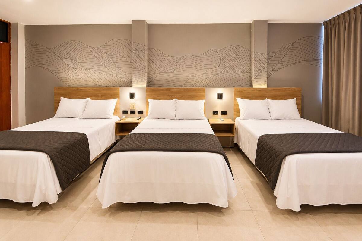 A hotel room with three beds, a wooden headboard, bedside tables with lamps, and a decorative wall design.