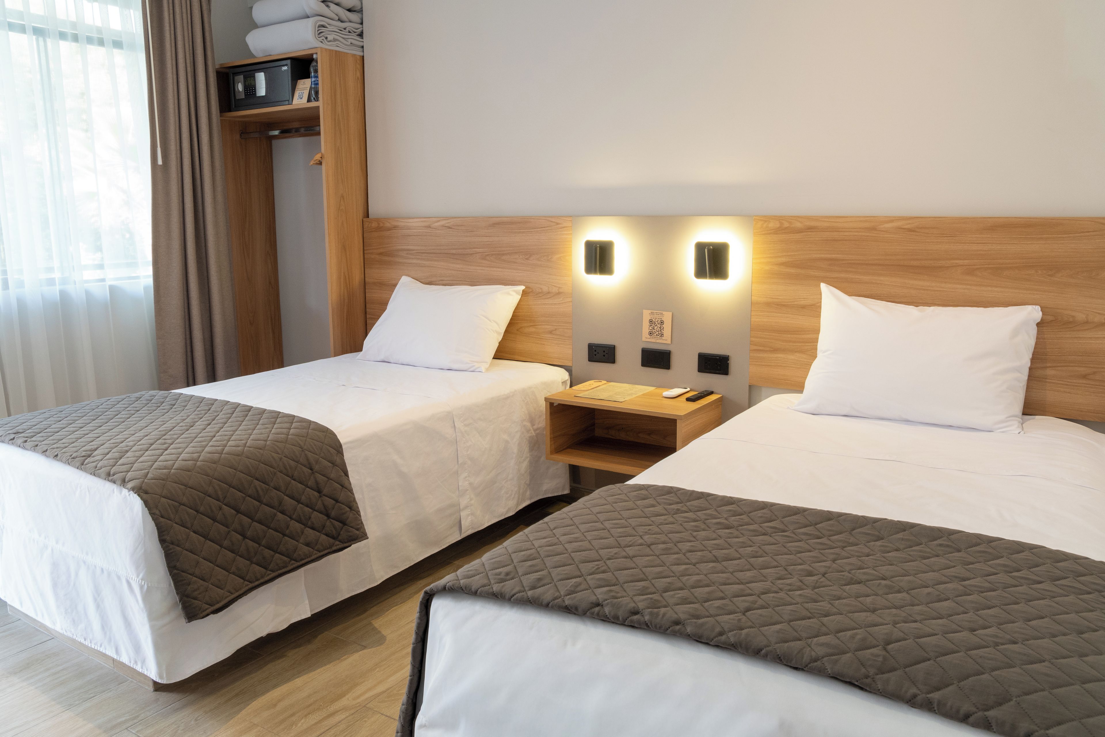 A hotel room with two single beds, a wooden headboard, bedside tables with lamps, and a window with curtains.