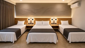 Habitación de hotel con tres camas individuales, cabecero de madera, luces fijadas a la pared y un diseño abstracto grande en la pared.