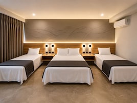 Habitación de hotel con tres camas individuales, cabecero de madera, luces fijadas a la pared y un diseño abstracto grande en la pared.