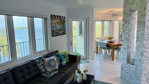 Un salon moderne avec un canapé foncé, un coin repas avec une table et des chaises en bois, et de grandes fenêtres offrant une vue sur la mer.