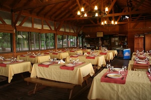 Restaurante com várias mesas preparadas para refeições, com vigas de madeira e grandes janelas.