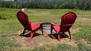 Two red Adirondack chairs facing a small fire pit in a grassy field with a mountain range in the background. - Fun Mountain Getaway with views and WIFI. Close to Mountains, Rivers and Lakes! (Wentworth)
