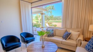 A room with a beige sofa, two blue armchairs, a wooden coffee table, and a view of a garden with a cactus and trees.
