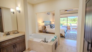 A modern bathroom with a large bathtub, a vanity with a mirror, and a view of a bedroom with a bed and a window.