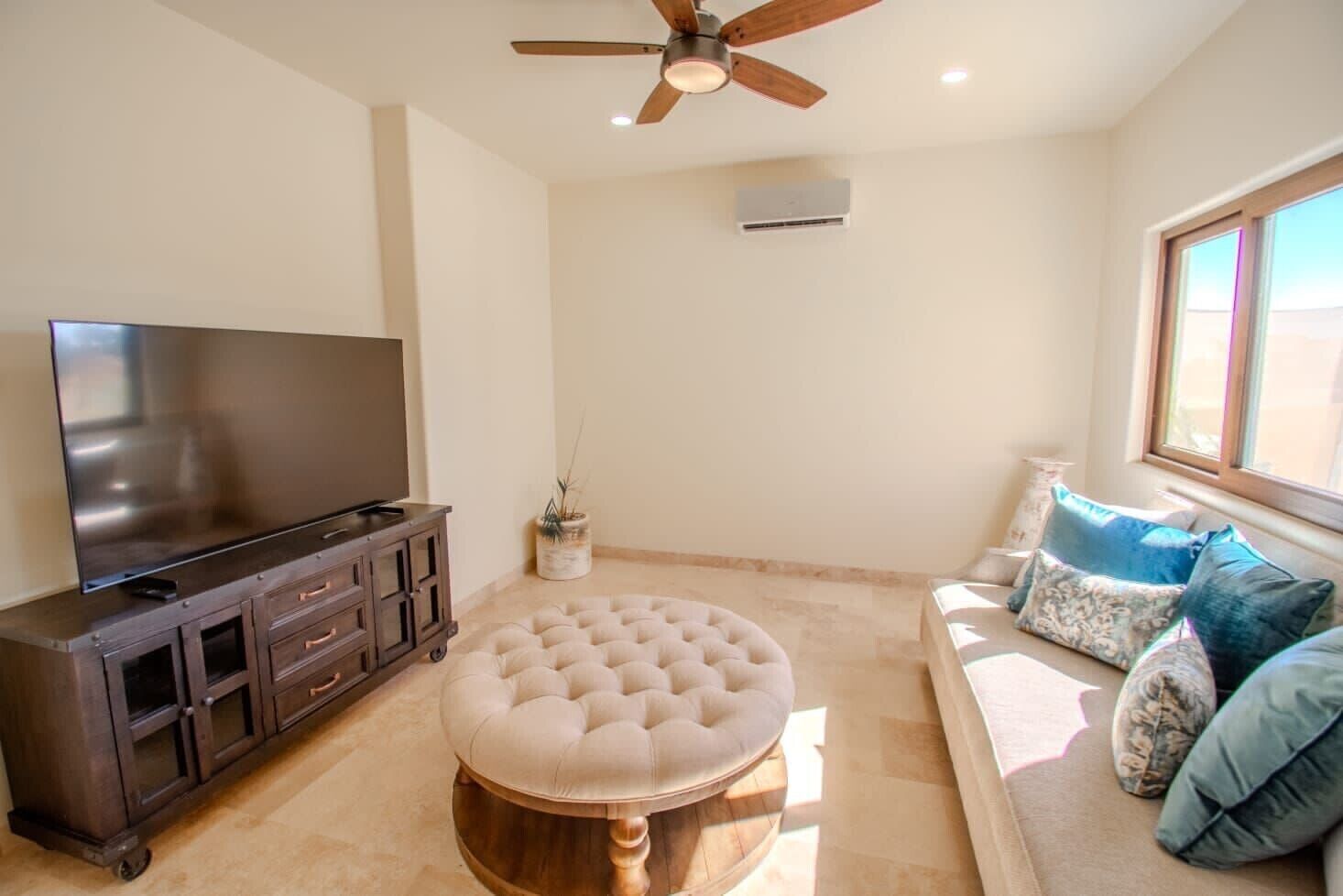 A living room with a flat-screen TV on a dark wooden cabinet, a round ottoman, a ceiling fan, and a window with blinds.
