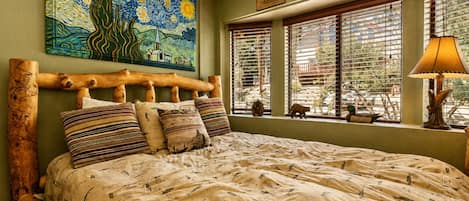 Una cama con cabecera de madera y ropa de cama estampada, un cuadro grande sobre la cama y ventanas con persianas.