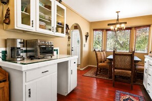 A kitchen with a coffee maker, oven, and cabinets. There is a dining area with a table and chairs. - Antelope Retreat (Big Bear City)