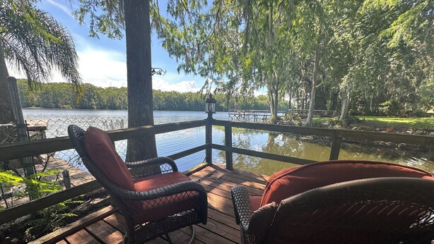 A wooden deck with wicker chairs and a lantern, overlooking a calm lake with trees and a dock in the background. - STUNNING WATERFRONT RETREAT!!! (Lake Panasoffkee)