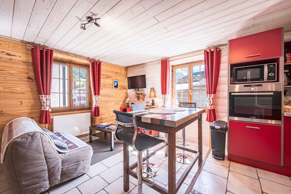 A cozy room with a wooden ceiling, a dining table with chairs, a sofa, a TV, and a red kitchen area with a microwave.