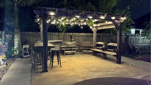An outdoor patio area with wooden benches and tables, string lights, and a wooden pergola. - Backyard Oasis in Brownsville! (Brownsville)