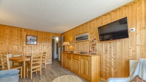 A wooden-paneled room with a dining table, chairs, a sofa, a TV mounted on the wall, and a kitchen area.
