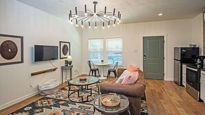 A modern living room with a brown leather sofa, a glass coffee table, and a patterned rug. There is a dining area with a small table and chairs, and a kitchen with a refrigerator and stove.