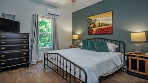 A bedroom with a bed, a dresser, a ceiling fan, a window with curtains, a painting on the wall, and two bedside lamps.