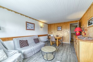 Une pièce chaleureuse avec un canapé, une petite table ronde, une table à manger avec des chaises et un coin cuisine agrémenté d’une lampe rouge.