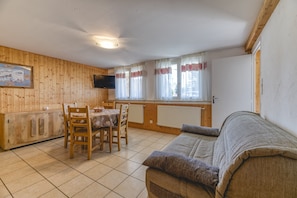 A room with a wooden interior, a dining table with chairs, a sofa, a TV mounted on the wall, and a window with curtains.