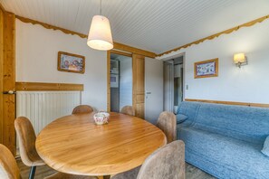 A dining area with a round wooden table, chairs, a blue sofa, and wall-mounted light fixtures. - Apartment 'Les Crocus 1-2' with Balcony and Wi-Fi (Huez)