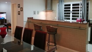 A modern kitchen with a glass island, two barstools, a dining table with chairs, a chandelier, and a television in the background.