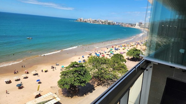 A beach with umbrellas and people, a clear sky, and a distant cityscape. - 04 BEDROOMS FACING THE SEA WITH AIR CONDITIONING IN TWO SUITES (Praia do Morro Guarapari)