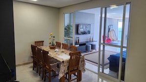 A dining area with a table set for six, a television mounted on the wall, and a sliding glass door leading to an outdoor seating area. - 04 BEDROOMS FACING THE SEA WITH AIR CONDITIONING IN TWO SUITES (Praia do Morro Guarapari)