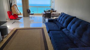 A modern living room with a blue sofa, a red chair, a dining area with a table and chairs, and a view of the ocean. - 04 BEDROOMS FACING THE SEA WITH AIR CONDITIONING IN TWO SUITES (Praia do Morro Guarapari)