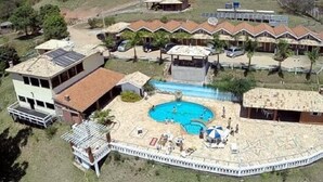 An aerial view of a resort with multiple buildings, a central swimming pool, and a fenced area with lounge chairs and umbrellas.