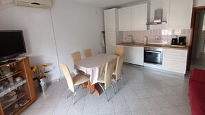 Une cuisine moderne avec des armoires blanches, une hotte de cuisinière en acier inoxydable et un four encastré. On y trouve une table à manger avec quatre chaises et un téléviseur posé sur un meuble en bois.