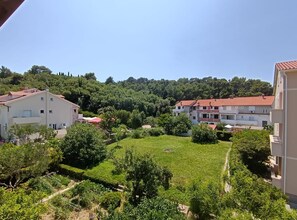 A residential area with multiple two-story buildings, a well-maintained grassy courtyard, and dense forested hills in the background. - Apartment Kurelić (Rab)
