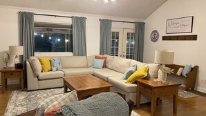 Een woonkamer met een modulaire bank, een houten salontafel en een bijzettafeltje met een lamp. Er is een raam met gordijnen, een plafondventilator en een welkomstbord aan de muur.