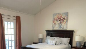 Een slaapkamer met een groot bed, houten vloer, plafondventilator, twee nachtkastjes met lampen, een raam met gordijnen en een bloemschilderij aan de muur.
