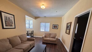 A living room with two sofas, a coffee table, and a dining table with chairs. There are windows with blinds, a ceiling light, and framed pictures on the walls.