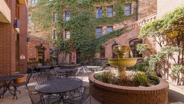 Un área de patio al aire libre con mesas y sillas redondas, una jardinera de ladrillo con una urna decorativa, y un edificio con paredes cubiertas de hiedra.