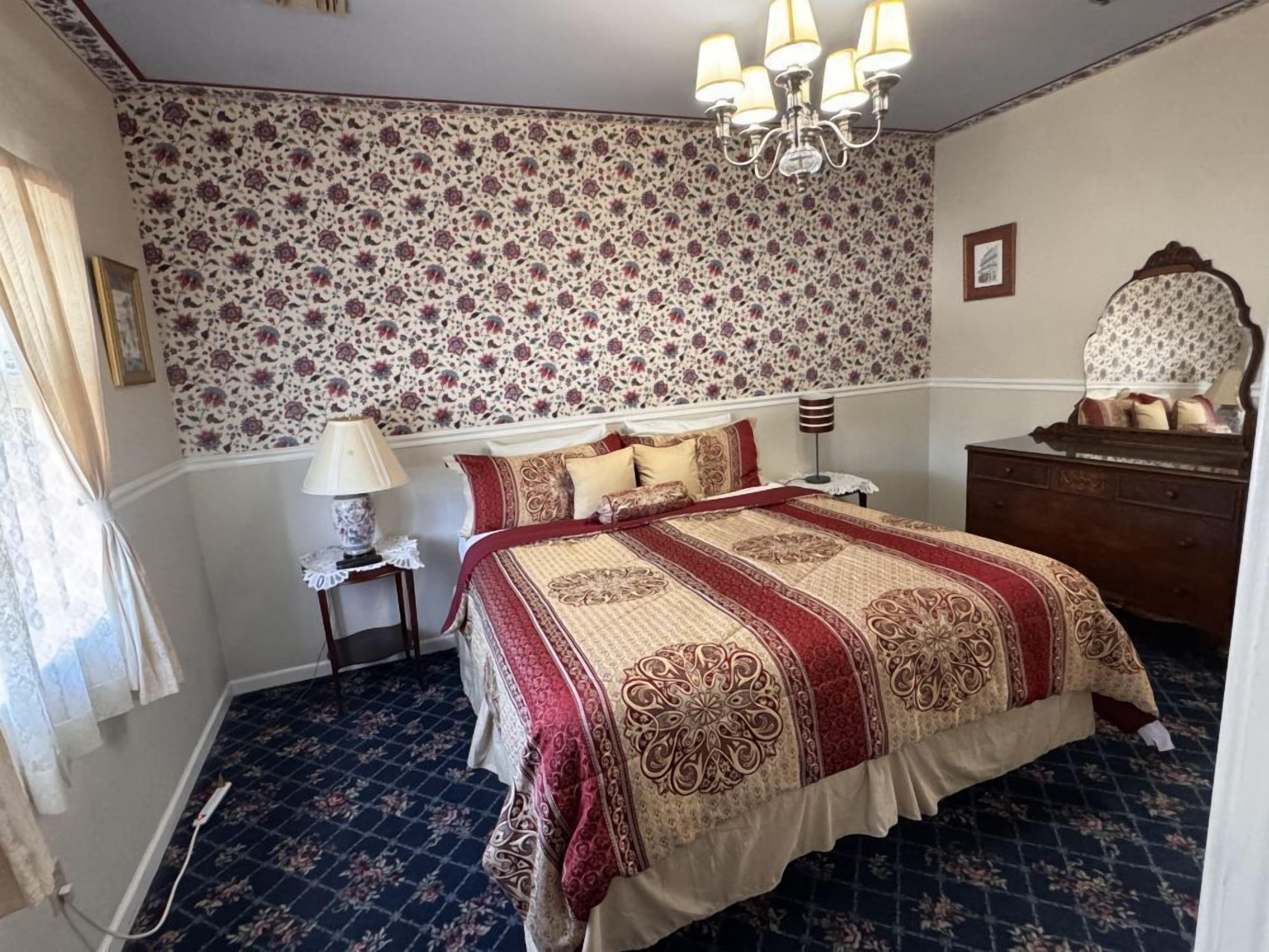 Una camera da letto con carta da parati a motivi floreali, un lampadario a quattro bracci, un comò in legno e una coperta con fantasia.