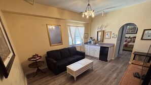 A compact living space with a black sofa, a white coffee table, a small side table, a kitchenette, and a dining area with a wooden table and chairs. - Charming Suite on Main Street (Placerville)