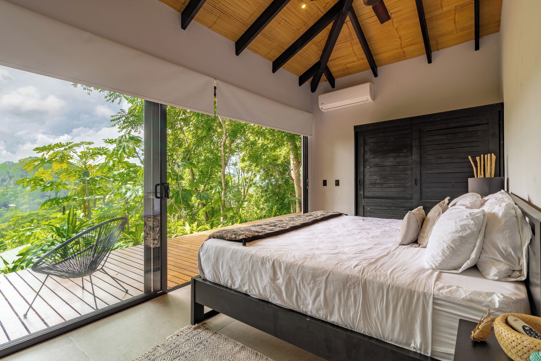 A bedroom with a large bed, a wooden ceiling, and a view of lush greenery outside.
