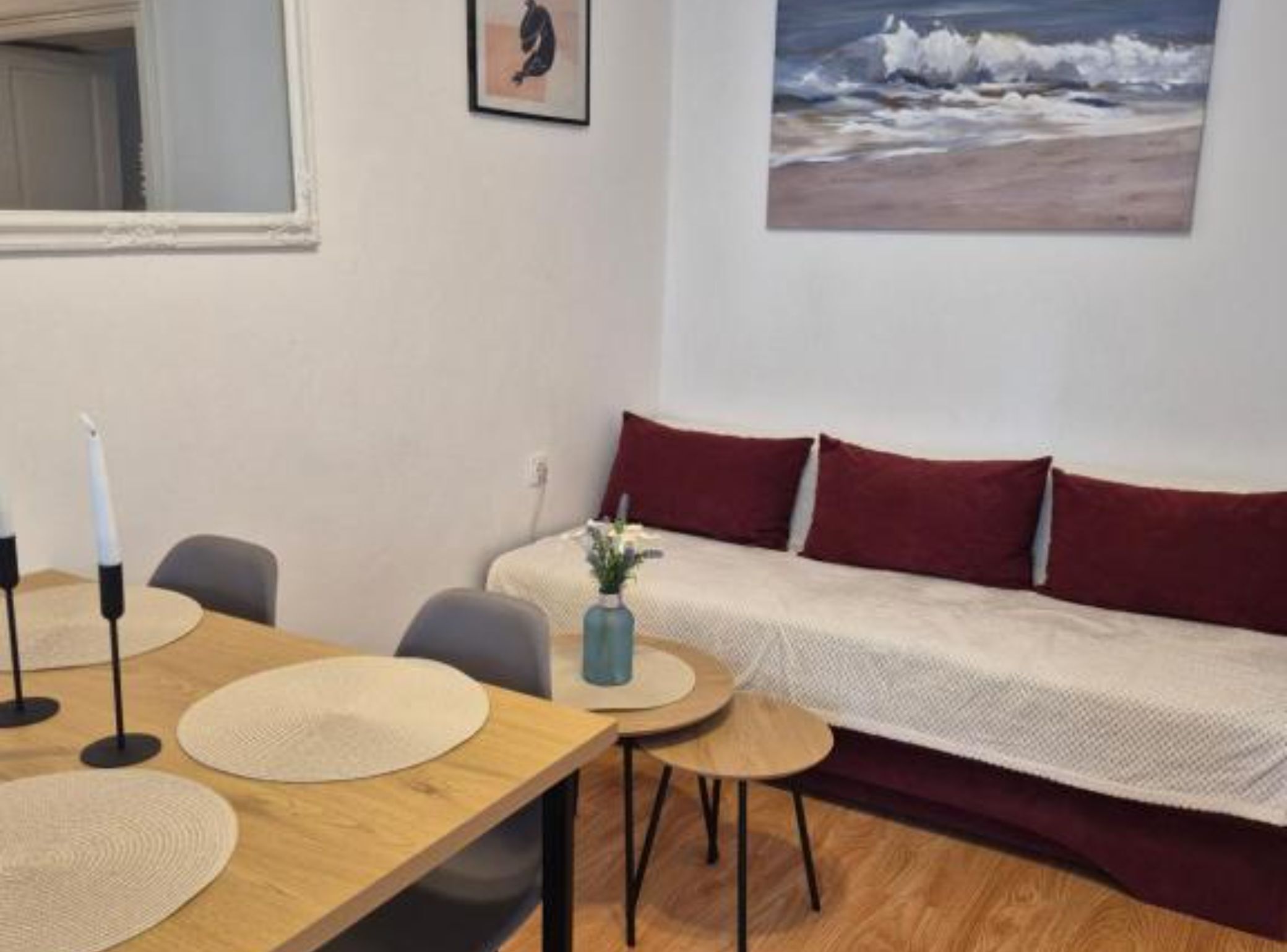 A cozy room with a wooden table, two chairs, a sofa with red cushions, and a painting of a beach scene.