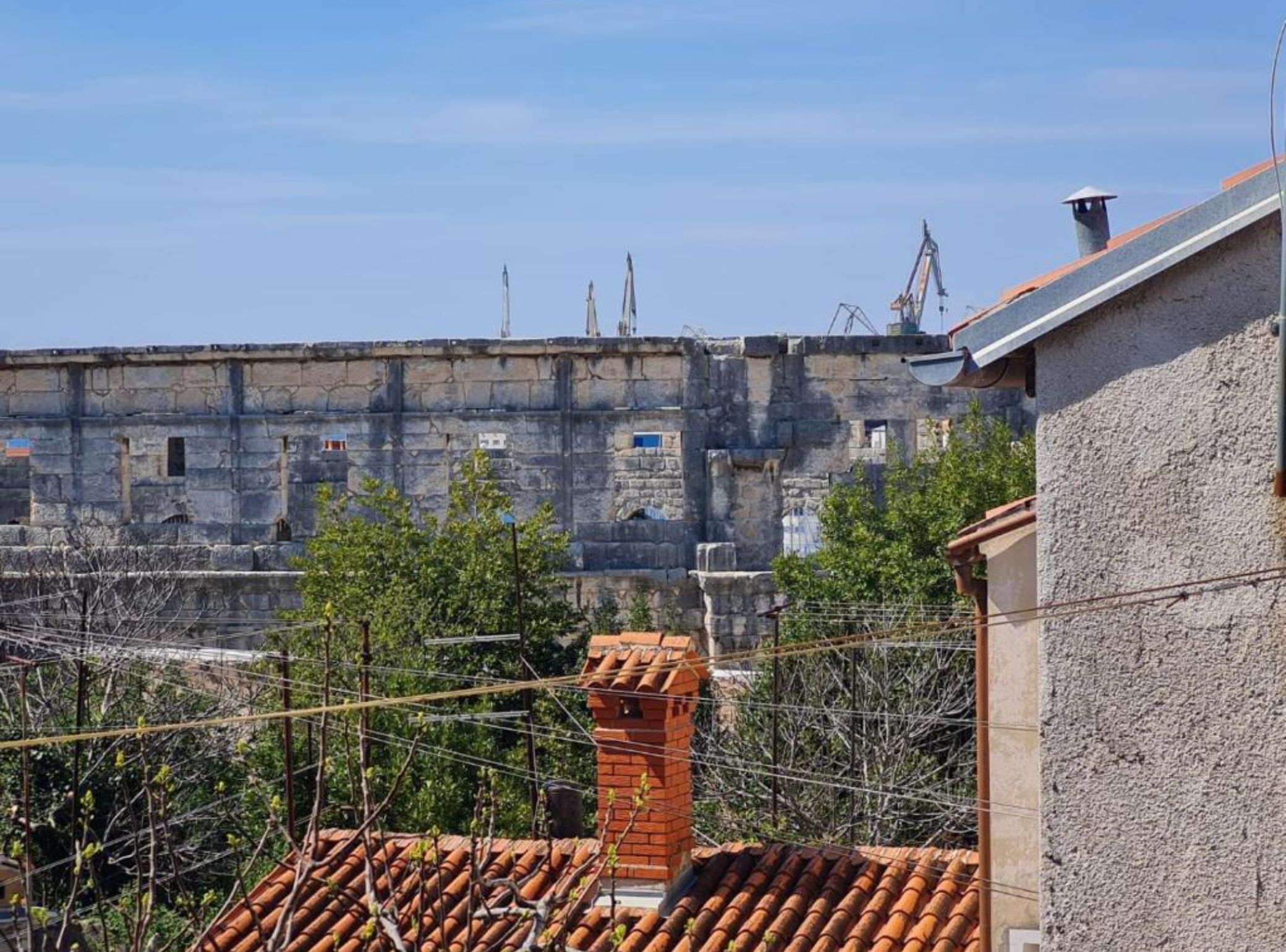 A view of a stone building with a tiled roof and a chimney, surrounded by greenery.