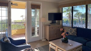 A living room with a TV, sofa, and chair, a balcony with a view, and a ceiling fan. - Eagle's Nest Gulf Front Unit, Closest to the Beach - Platinum Week 11 (Marco Island)