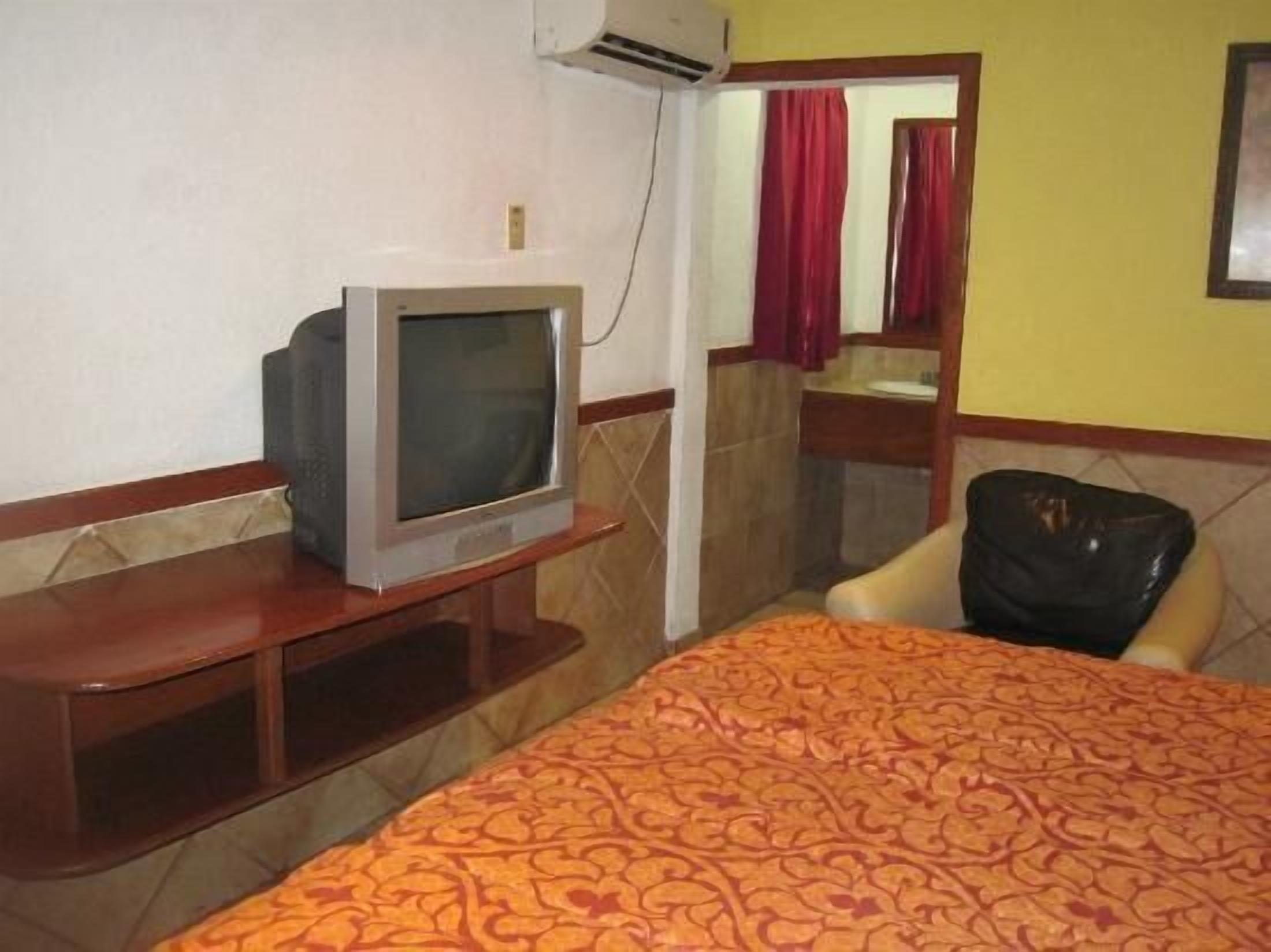 A hotel room with a bed, a television on a wooden stand, a chair, and a bathroom visible through a glass door.