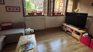 Ein Wohnzimmer mit Holzboden, einer Couch, einem Couchtisch mit einem Buch, einem Fernseher auf einem Holzschrank und einem Fenster mit Blumenkästen.