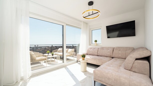 A spacious living room with a large window, a beige sofa, a round coffee table, and a flat-screen TV mounted on the wall.