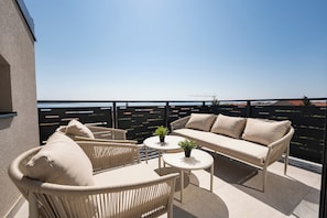 A balcony with wicker furniture, a sofa, and two small tables, overlooking a body of water and a construction site. - Dominik (Mandre)