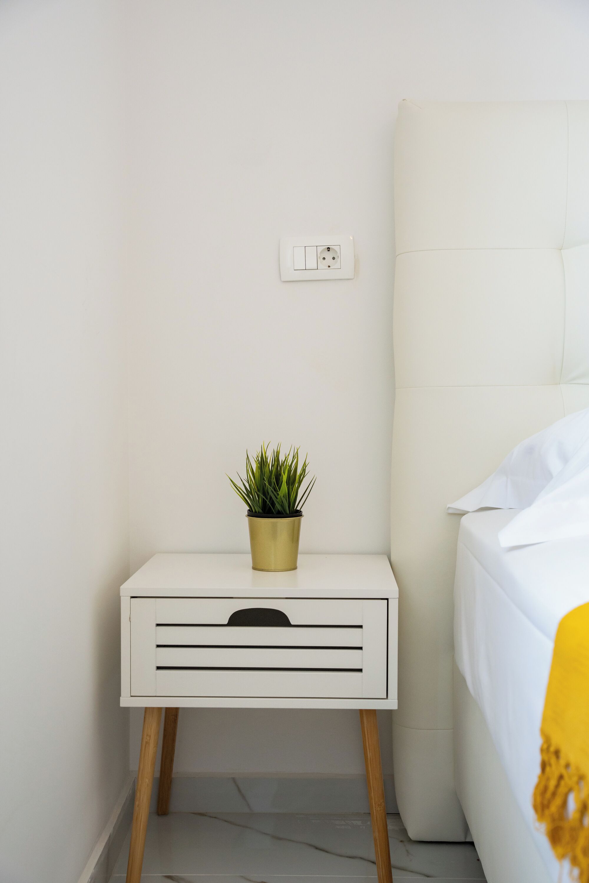 A white bedside table with a gold pot of green plant, a white bed with a white pillow, and a white wall with a power outlet.