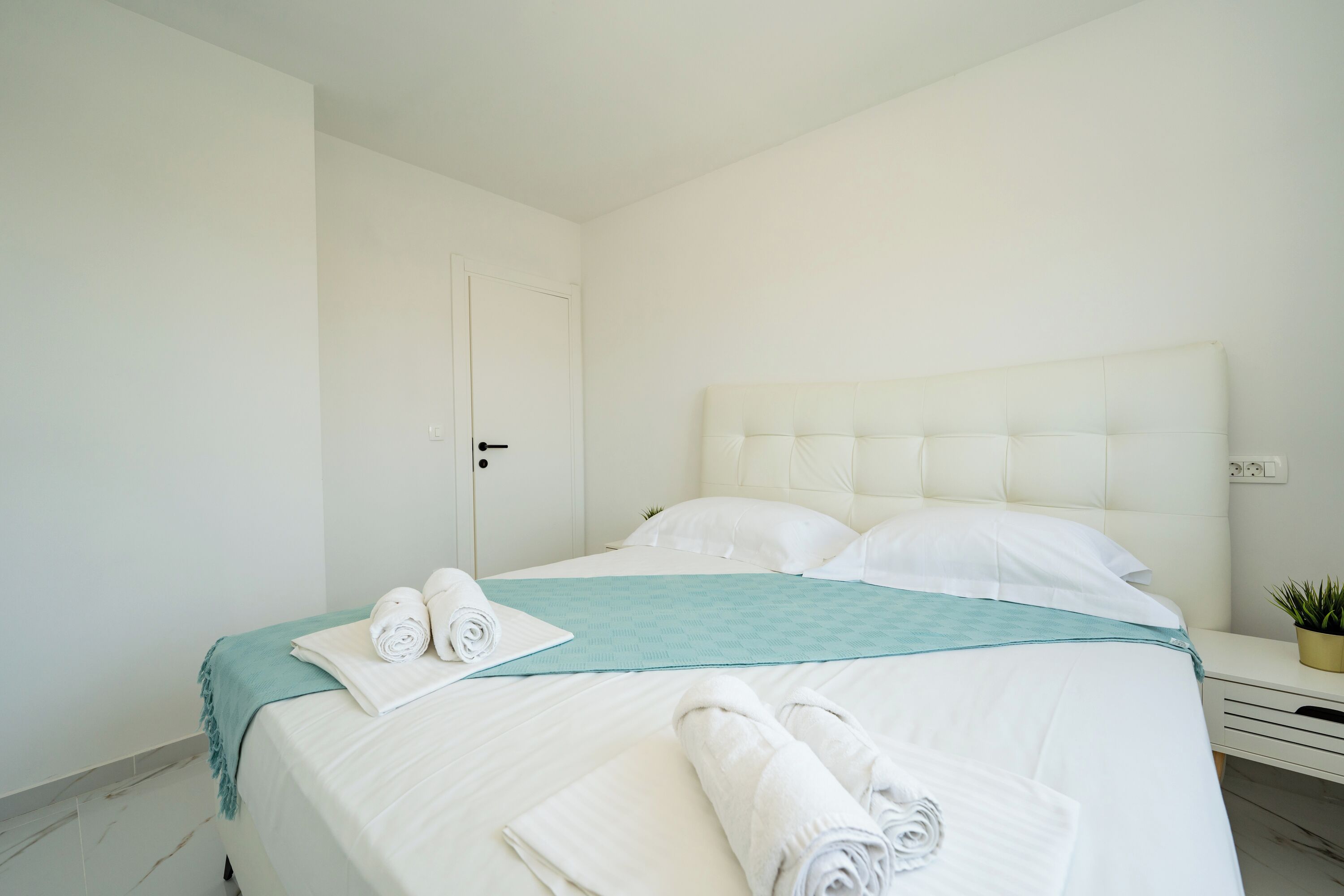 A neatly made bed with white linens and a teal throw, a bedside table with a plant, and a door with a handle.