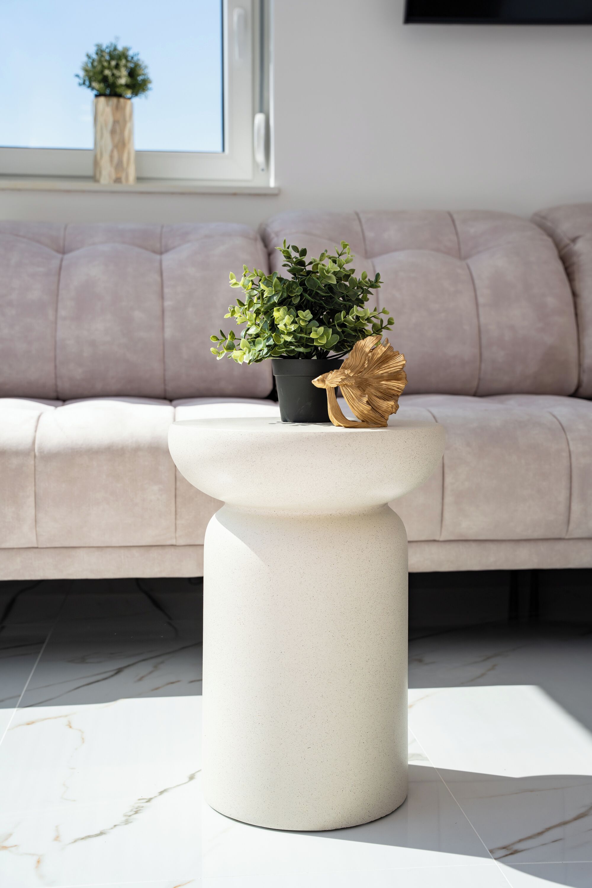 A modern living room with a light-colored sofa, a white pedestal table with a potted plant and a decorative item, and a marble floor.
