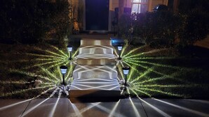 Un chemin menant à l’entrée d’un bâtiment, avec une porte sombre, flanqué de lanternes bien éclairées et de haies taillées.