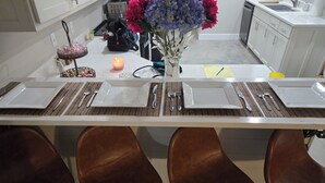 Une cuisine avec un long comptoir, quatre chaises marron, un vase rempli de fleurs et une bougie.