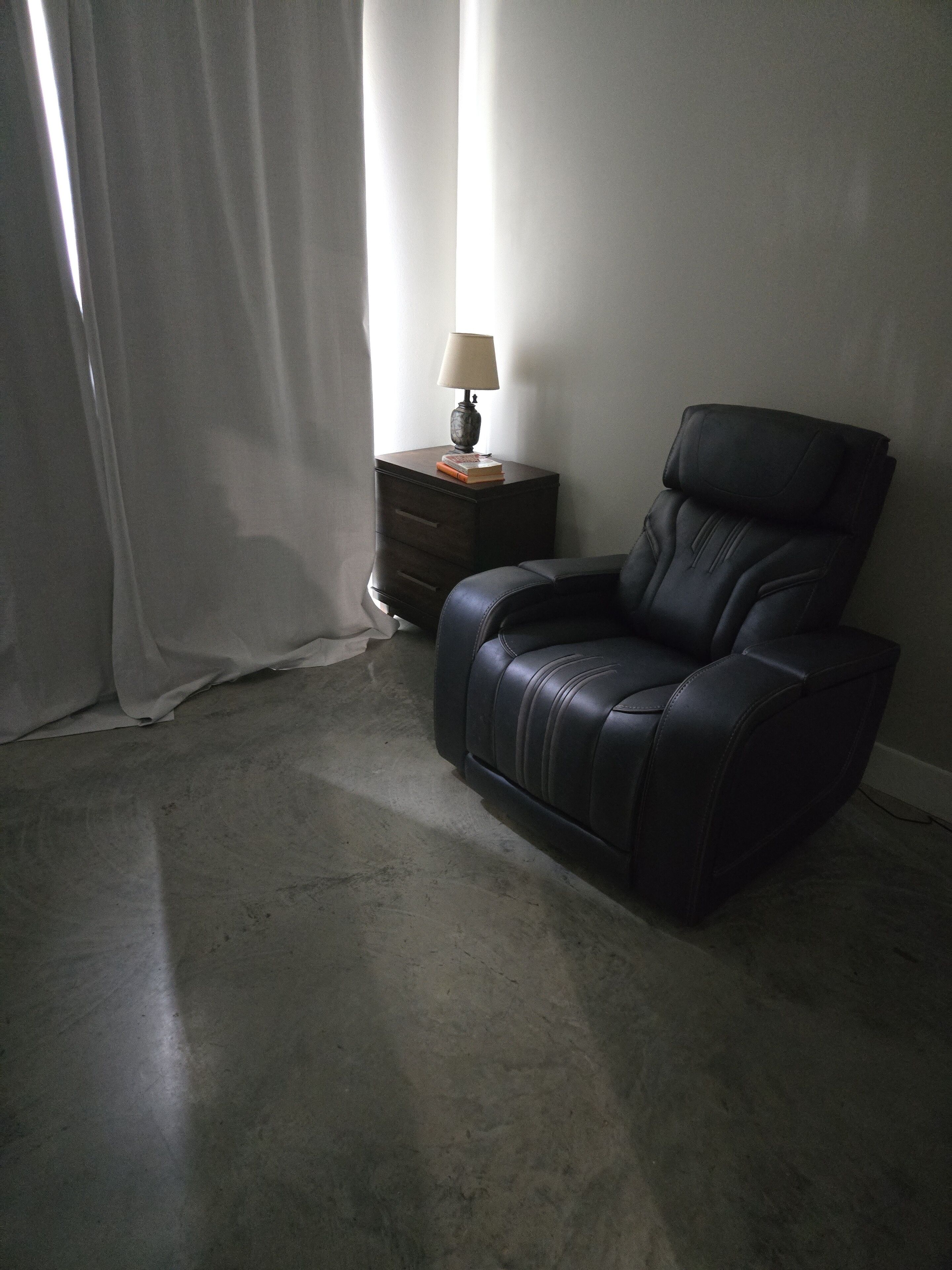 A dark leather chair, a small wooden nightstand with a lamp, and a plain wall.