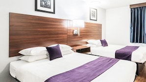 A hotel room with two beds, each with white bedding and purple accents, a wooden headboard, a bedside table, and a wall-mounted lamp. - Knights Inn Pilot Mountain (Pilot Mountain)